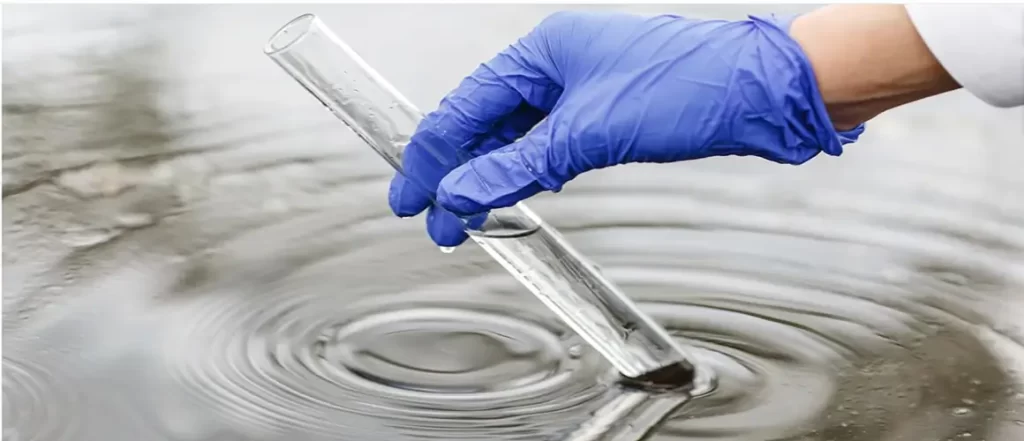 A gloved hand holds a test tube collecting water from a pond, creating ripples on the surface.